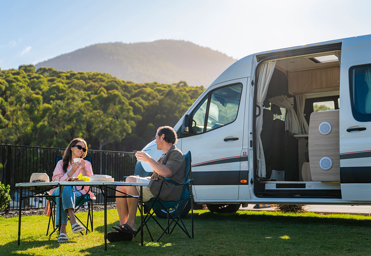 Couple enjoying a meal on their outdoor camping setup in from of their winnebago campervan with the side sliding foor open, outdoors sunny day with a forest and mountains in the backgroud.